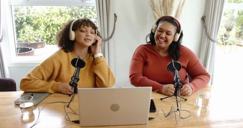 Two friends recording lively podcast in bright home studio with microphones and laptop