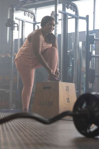 African american woman tying sneaker on numbered plyo box in gym, wearing pink activewear