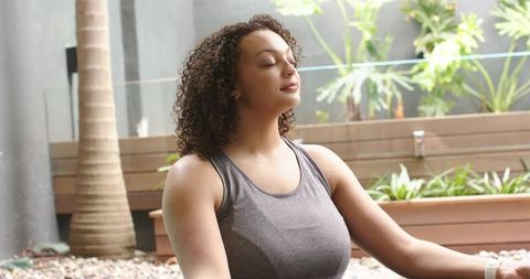 African american woman meditating on terrace with pebble patio, wooden planters and greenery