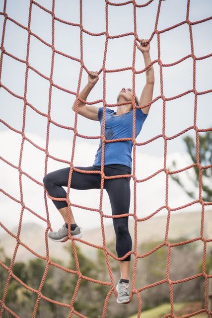 Fit woman climbing a net during an outdoor obstacle course in a boot camp. Ideal for use in fitness, training, and outdoor adventure themes. Perfect for promoting exercise programs, boot camps, and athletic events.