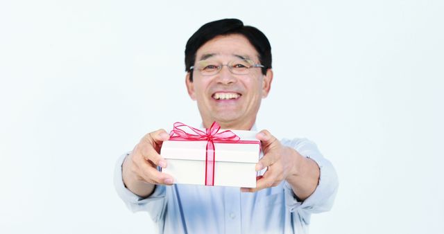 An older man wearing glasses is smiling and holding out a white gift box wrapped with a red ribbon. The background is clean and minimal, enhancing the focus on the man and the gift. Ideal for use in themes related to celebrations, generosity, birthdays, holidays, or special occasions. Can be used in promotional materials, greeting cards, advertisements, or social media posts.