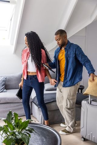 Young black couple packing luggage in bright loft apartment holding straw hat and tablets