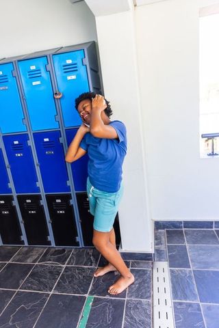 African american teen leaning by blue lockers adjusting hair barefoot in school corridor