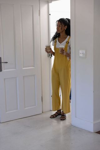 African american woman entering home wearing mustard overalls holding two coffee cups