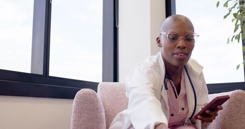 African female doctor using smartphone while reaching in clinic armchair during shift