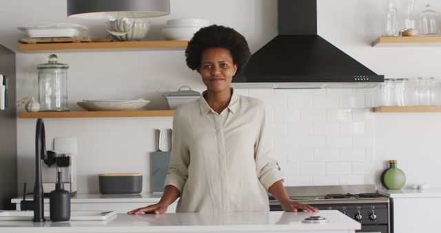 Woman standing in a modern kitchen, smiling confidently. The kitchen features natural light, minimalist design, and organized shelves with glassware and dishes. Perfect for themes of modern living, home decor, kitchen design, everyday life, and domestic scenes.