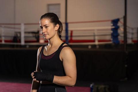 Female boxer adjusting hand wraps near boxing ring, focused athlete training in gym