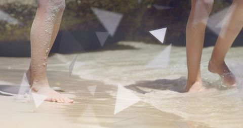 Young girls wading barefoot along sunlit sandy shore, sand-dusted legs, shallow surf foam