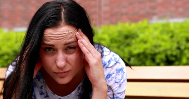 Image depicts a young woman with dark hair holding her head in pain while sitting on a bench. She appears stressed and worried, suggesting a headache or other discomfort. Suitable for use in articles or advertisements related to mental health, stress management, physical pain, or wellness products.