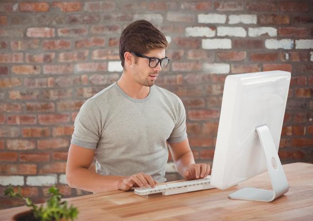 Man is working on a desktop computer in a modern office with a brick wall background. He is wearing glasses and a casual grey shirt. This image can be used to illustrate office work, technology usage, working professionals, and modern office environments.