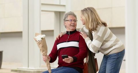 Mature couple sharing flowers and conversation on urban bench, man holding smartphone