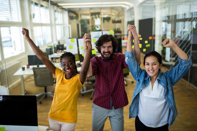 Diverse group of graphic designers holding hands and cheering in a modern office environment. Ideal for use in articles or advertisements about teamwork, collaboration, workplace success, and creative industries. Can be used to illustrate concepts of unity, motivation, and professional achievement.