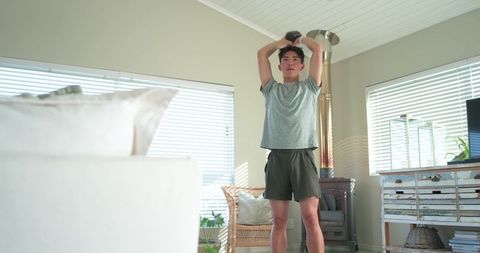 Young man lifting dumbbell overhead during home workout in bright living room