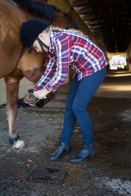Young girl wearing plaid shirt and helmet grooming a horse in a stable. Ideal for themes related to equestrian activities, animal care, rural lifestyle, and youth involvement in horse riding. Useful for websites, blogs, and promotional materials focused on horse care, farm life, and equestrian sports.