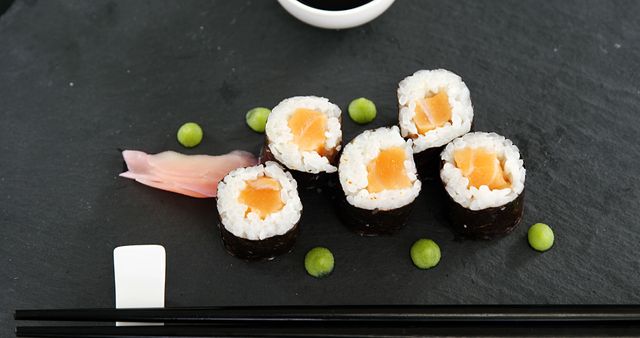 A delicious arrangement of salmon sushi rolls is presented on a slate board, with soy sauce, wasabi, and pickled ginger, with copy space. Sushi is a popular Japanese dish that has gained international acclaim for its delicate flavors and artistic presentation.
