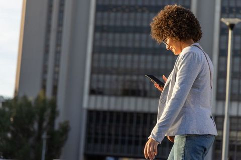 Urban professional walking, checking smartphone near office tower during golden hour
