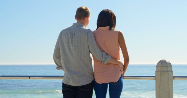 A young Caucasian couple stands close together, looking out at the sea, with copy space. Their relaxed posture and the serene ocean backdrop suggest a moment of peaceful contemplation or a romantic getaway.