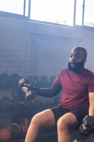 African american man sitting on bench lifting dumbbells in industrial gym strength focus