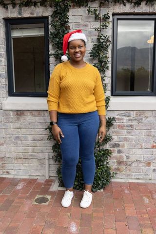 African american woman standing in santa hat and mustard sweater smiling by brick wall