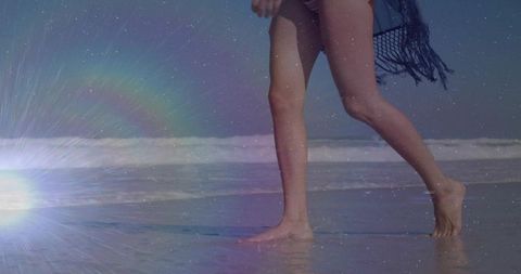 Barefoot woman walking on wet sand with sarong at sunlit beach, ocean waves and lens flare