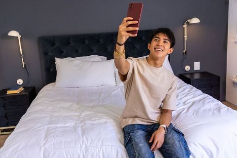 Smiling asian man sitting on bed taking selfie with red smartphone in modern bedroom
