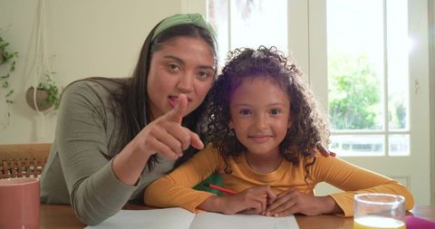 Mother guiding daughter writing homework at sunlit kitchen table, teaching and encouraging