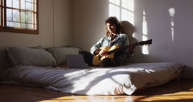 Relaxed biracial woman sitting on bed playing acoustic guitar in sunny cottage bedroom. simple living in off the grid rural home.