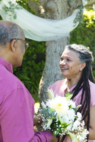 Senior couple exchanging white bouquet and smiling under draped tree in garden ceremony