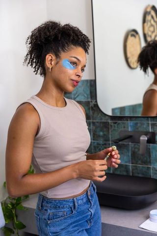Woman applying blue hydrogel eye patches at modern bathroom vanity, skincare routine