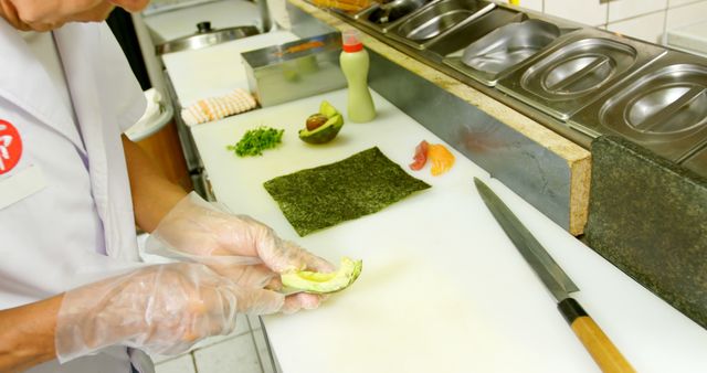 Chef is preparing sushi using fresh ingredients in a professional kitchen. The workspace is clean and organized, showcasing essential sushi-making tools and ingredients like nori, avocado, and fish. This image can be used in articles, advertisements, or websites related to culinary arts, Japanese cuisine, or professional cooking environments.