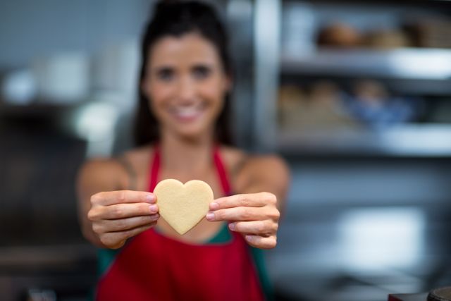 Female baker smiling and holding a heart-shaped cookie in a bakery. Ideal for use in advertisements for bakeries, baking classes, or Valentine's Day promotions. Can also be used in articles about baking, food preparation, or small business success stories.