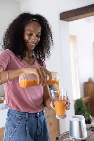 African american woman pouring fresh carrot smoothie from blender into glass at home