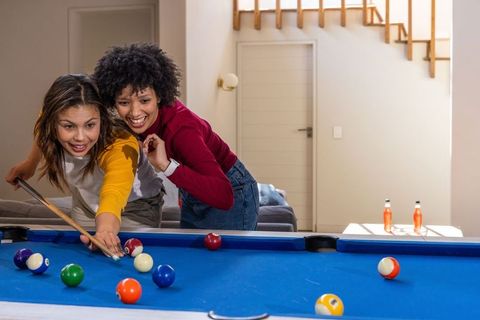 Friends playing pool on blue table in cozy living room, diverse women aiming and laughing