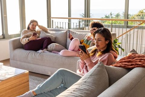 Multicultural women lounging on sofa scrolling smartphones in bright coastal living room