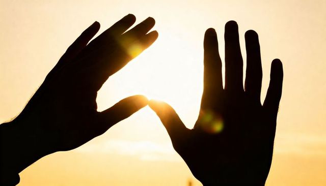 Silhouetted hands framing rising sun at golden hour with warm lens flare and backlight horizon