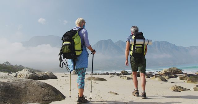 Senior couple with backpacks hiking along a scenic rocky beach on a misty morning. The man and woman enjoying the natural beauty of the coast, suggesting a healthy and active retirement lifestyle filled with outdoor adventures and exploration. Perfect for travel advertisements, nature blogs, and health and wellness promotions targeting older adults.
