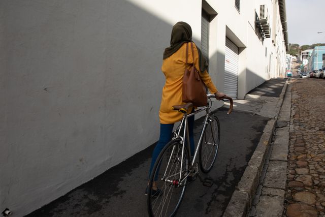 Rear view of biracial woman wearing hijab and yellow jumper out and about on the go in the city, walking with bike. Commuter modern lifestyle.