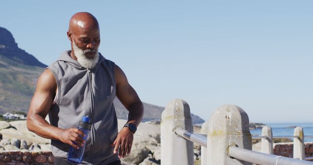 Senior man taking a break during outdoor exercise by the beach, holding a water bottle. Ideal for promoting senior fitness, healthy living, and active retirement lifestyle.