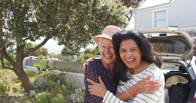 Two senior women share a joyful embrace outdoors, both smiling brightly. The scene suggests deep friendship and happiness, surrounded by a garden with trees and a house in the background. Ideal for use in advertisements, articles, and promotional materials highlighting themes of friendship, aging gracefully, and outdoor activities for seniors.
