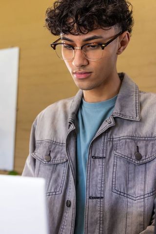 Asian young man working on laptop wearing tortoiseshell glasses and denim jacket
