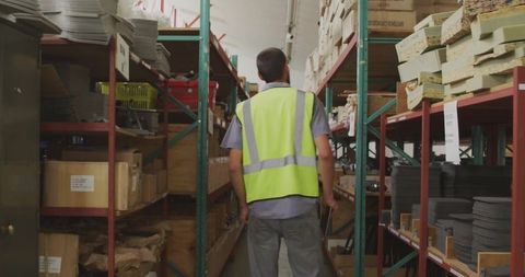 Warehouse worker inspecting inventory in aisle wearing hi-vis vest and holding clipboard