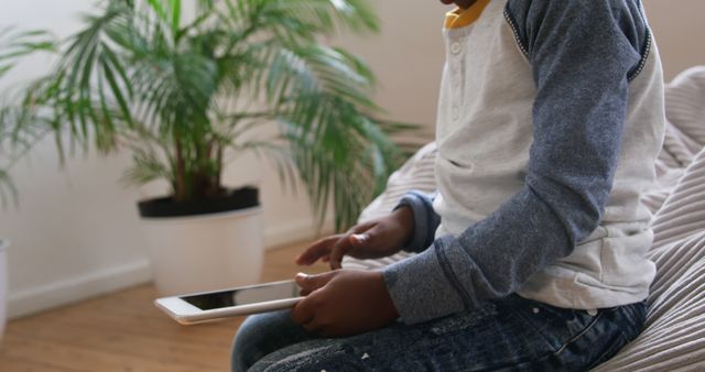 Young boy in casual clothes using tablet at home, near potted plants. Ideal for content about children's activities, home learning, digital interaction, relaxing at home, and modern technology usage for kids.