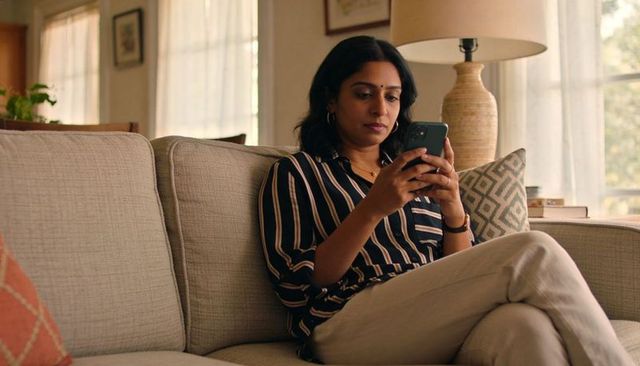 Indian woman sitting on sofa using smartphone in sunlit living room for modern lifestyle