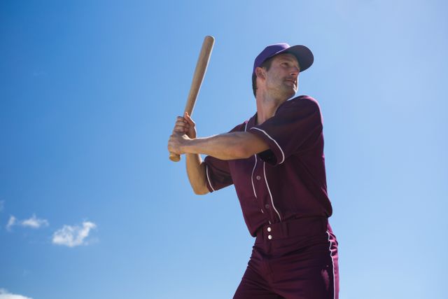 Young baseball player in uniform holding bat, ready to swing, against clear blue sky on a sunny day. Ideal for sports-related content, athletic promotions, and outdoor activity themes.