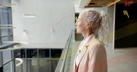 Mature professional woman in pink blazer standing at glass railing in modern atrium