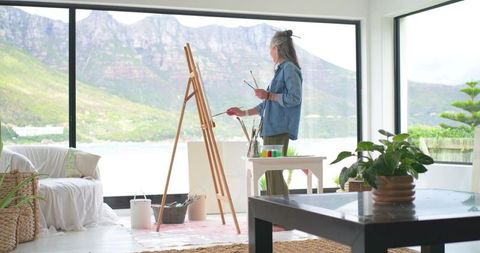 Senior woman painting at easel in bright lakeside studio with mountain view