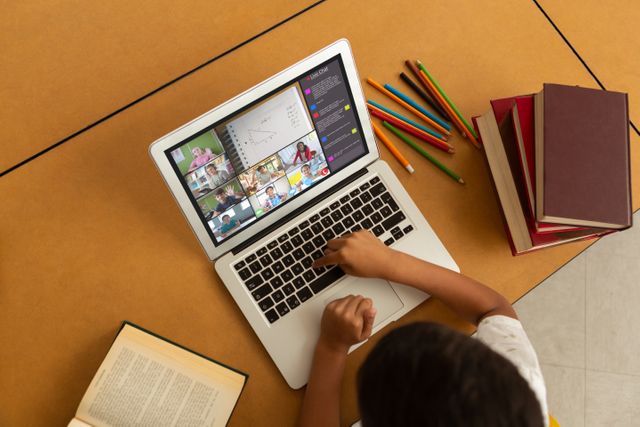 Child engaged in an online class using a laptop, surrounded by books and colored pencils. The child is learning remotely, participating in virtual education. Ideal for illustrating remote learning, online classes, educational technology, home study environments, and virtual school settings.