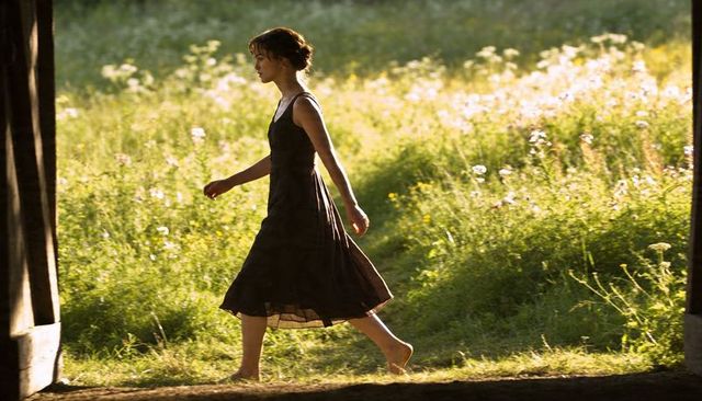 Barefoot woman stepping through timber doorway into sunlit wildflower meadow, backlit silhouette