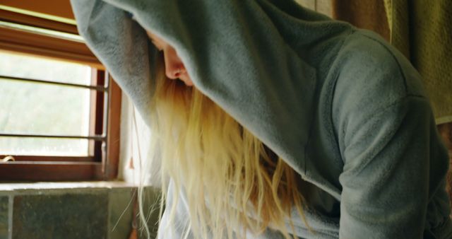 Woman wearing grey hoodie leaning near window. Blonde hair falling forward obscuring face. Natural light illuminating scene. Person appearing reflective and relaxed. Suitable for themes of contemplation, everyday lifestyle, natural beauty, stillness, and introspection.