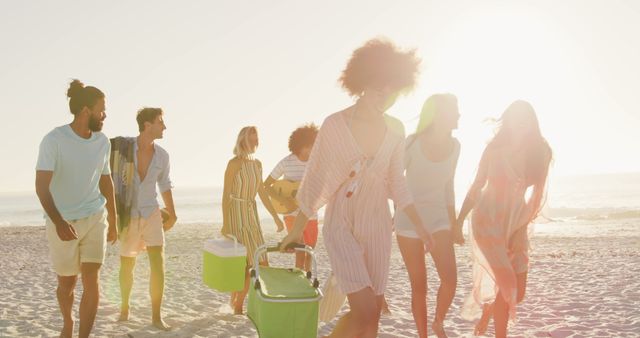 Happy diverse friends walking on beach with drink coolers, bags and beach blanket at sundown. Summer, free time, friendship, relaxation and vacations.